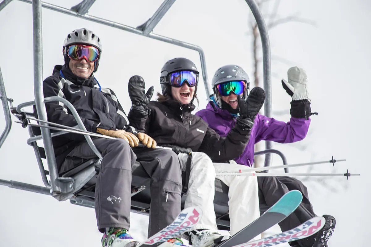 Three skiers ride a chairlift high above a snowy landscape, clad in vibrant ski gear with spectacular mountain views all around. The composition conveys the camaraderie and joy of a ski day, perfectly capturing the allure of Telluride’s winter sports and social atmosphere.