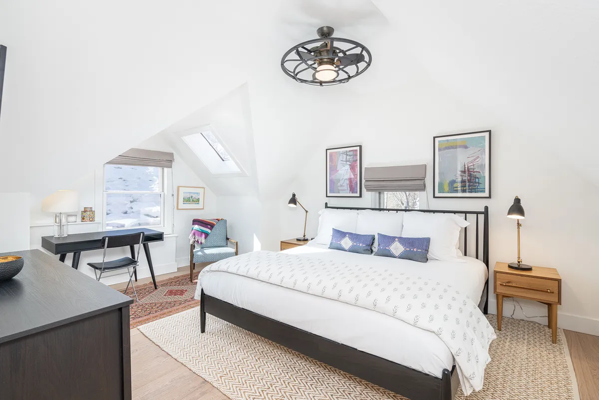 This image shows a bedroom with a bed adorned with decorative pillows, artwork on the walls, a desk and chair, a window allowing natural light to fill the space, and a rug on the floor. The arrangement is serene and welcoming, ideal for restful sleep or a quiet workspace.