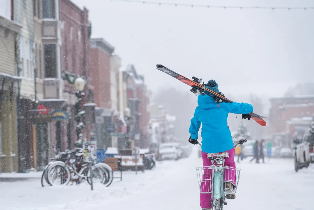 A snow-covered street winds through Telluride’s historic downtown, lined with charming 19th-century buildings. A person pedals a bicycle fitted with skis, blending winter adventure with everyday mountain-town life. The bright facades of the storefronts and snow-dusted roofs evoke a sense of place and heritage.