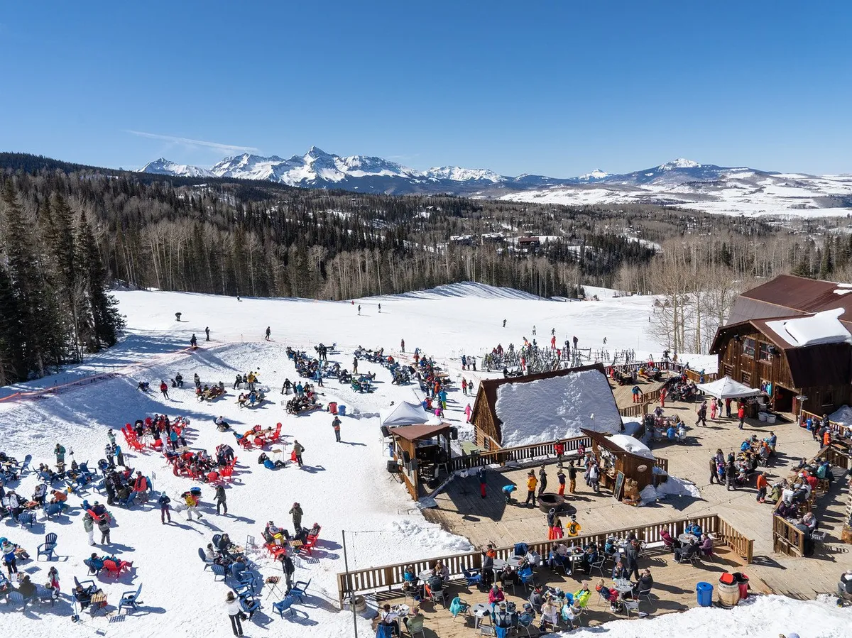 A bustling outdoor scene at the base of the ski slopes, with groups of people gathered around an outdoor seating area, set against a backdrop of majestic mountains and a ski lodge. The image radiates a joyful, communal energy reflective of Telluride’s downtown vibrancy.