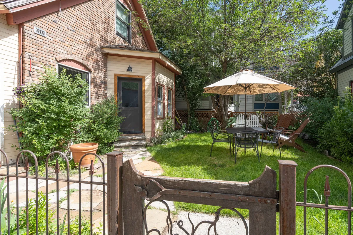A private garden courtyard with a seating area beneath an umbrella, surrounded by lush greenery and the house's inviting facade. The space is set up for outdoor dining, relaxing, and safe play, evoking a tranquil retreat in summer months.