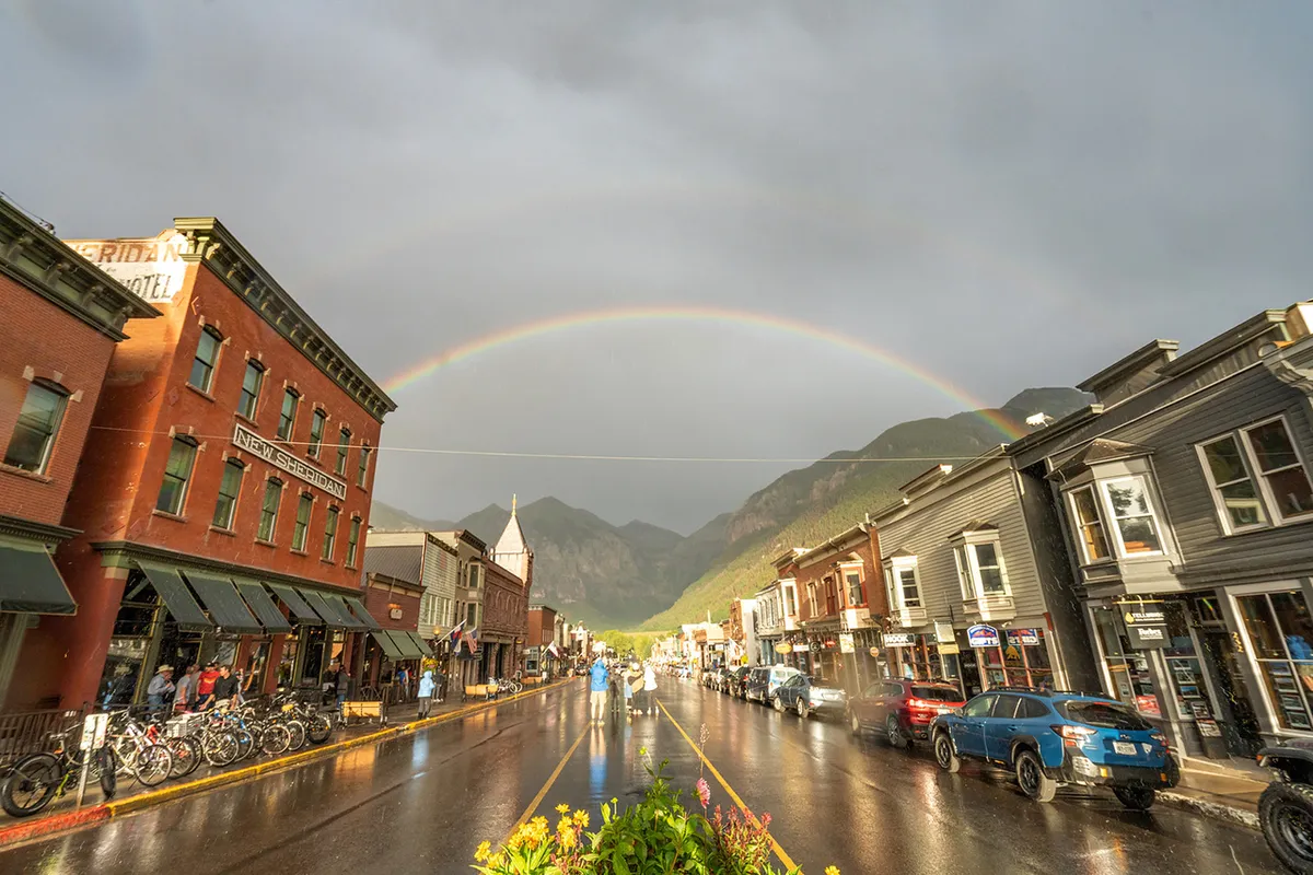 A picturesque Telluride street scene featuring historic buildings, a rainbow, wet pavement, and mountains in the background. Bicycles are parked nearby, suggesting accessibility and a vibrant local community spirit. The image conveys the charm and walkability of the area, as described in the narrative.