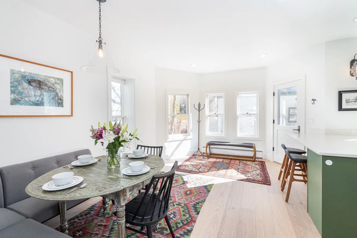 A dining area with a table adorned by a floral centerpiece, surrounded by large windows streaming with natural light, and complemented by a colorful area rug. The setup is intimate and inviting, perfect for group meals and lively conversations.