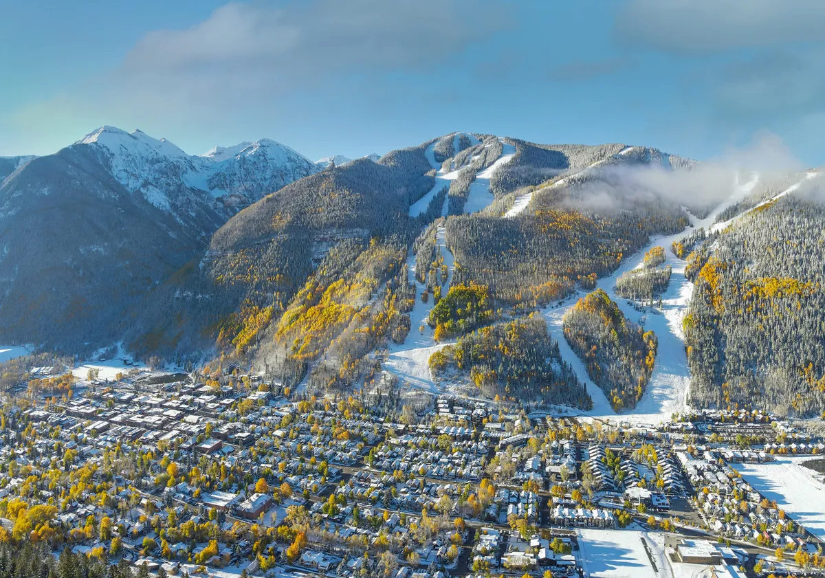 A sweeping view of Telluride from above, showing snow-capped mountains, colorful autumn trees, ski slopes, and the quaint town nestled below. The image conveys the proximity of adventure, scenic beauty, and the unique setting of the town.
