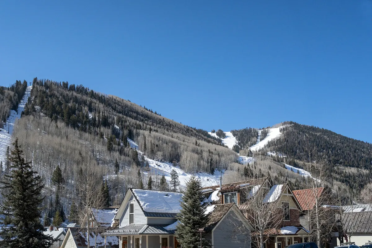 The image reveals a picturesque view of Telluride’s snow-covered mountains rising above a collection of quaint houses and evergreen trees. The small-town neighborhood ambiance is enhanced by the cozy building exteriors nestled in the scenic landscape, inviting exploration and discovery.