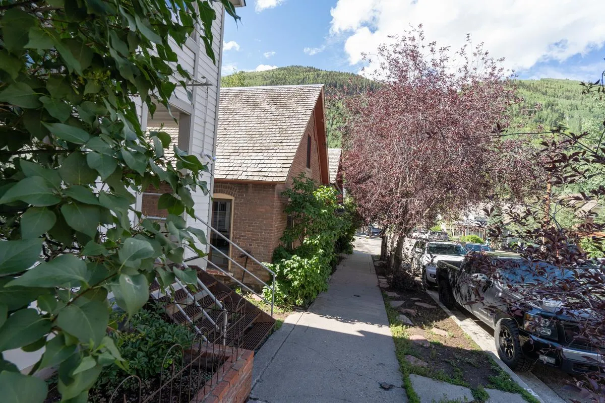 A sunlit brick building set amidst green foliage, with a pathway leading toward it and majestic mountains rising in the background. The scene suggests a vibrant, walkable neighborhood with natural beauty and convenient proximity to local attractions.