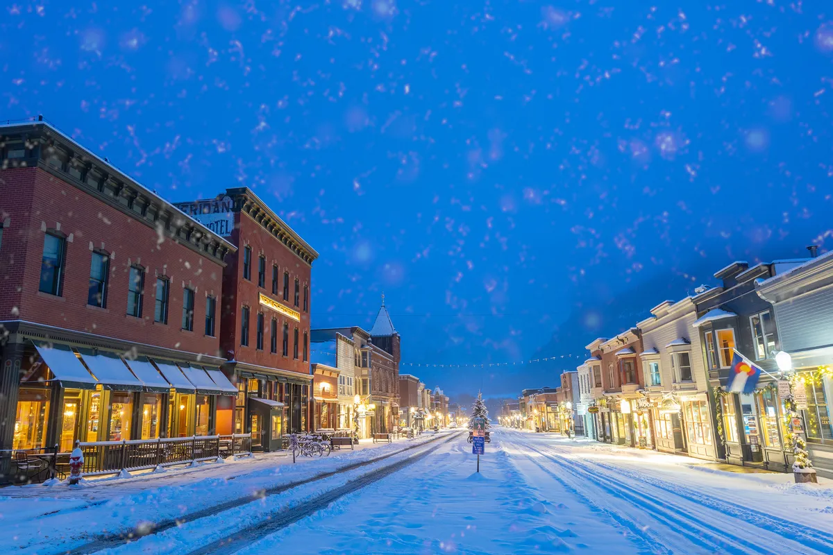 A picturesque, snow-covered street in Telluride featuring historic buildings, decorated trees adorned with lights, and warmly glowing streetlights. The scene captures the town’s vibrant yet cozy atmosphere, with people strolling along the sidewalks, suggesting easy access to shops, dining, and nightlife.