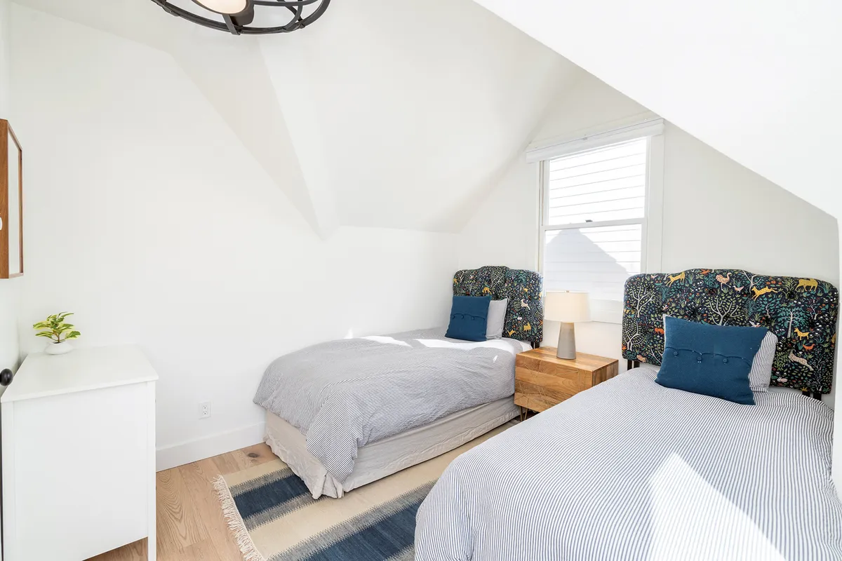 This image features a cheerful bedroom with two beds topped by colorful headboards, a stylish light fixture, a bedside lamp, and a textured rug. The mood is playful yet comfortable, suitable for both kids and adults.
