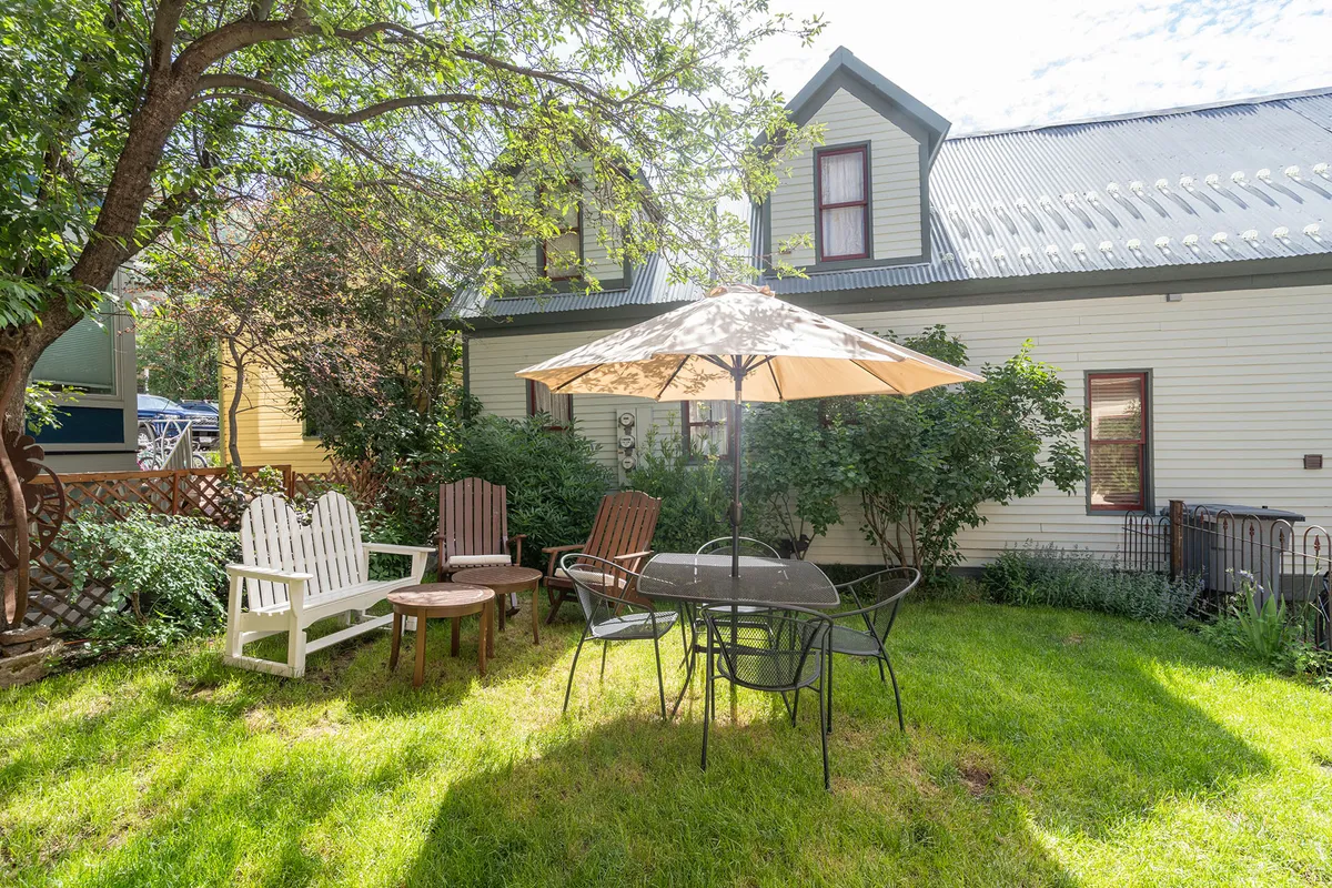 A charming outdoor patio with a table and chairs shaded by an umbrella, surrounded by lush greenery and the facade of the house visible in the background. The area is sunlit, with space for gatherings or quiet relaxation, evoking a sense of summer leisure and outdoor enjoyment.