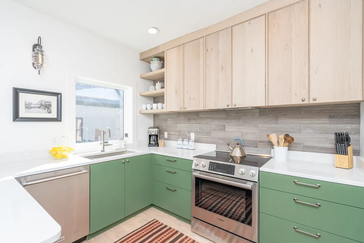A bright, cheerful kitchen featuring green cabinetry, sleek stainless steel appliances, a sunlit window, and open shelving displaying dishware. The space is perfect for baking, cooking, and socializing, echoing a modern and inviting vibe.