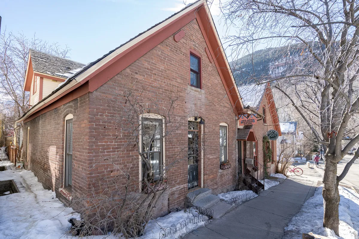 A charming exterior shot of the Twin Bricks building, featuring its distinctive brick facade, front steps, and mature tree branches. The image captures the welcoming presence of the home within its walkable, lively neighborhood setting.