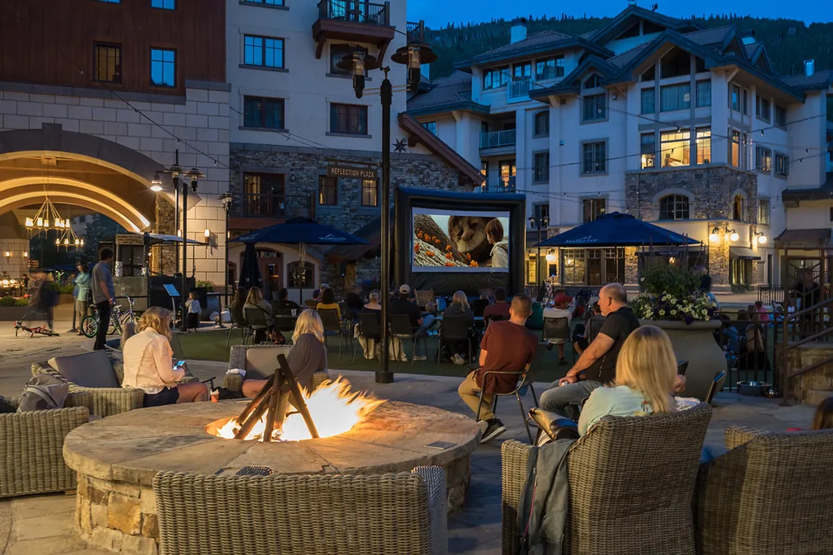 An inviting outdoor setting featuring a fire pit, a cozy seating area, and an outdoor movie screen, all under the open sky. The mood is festive and warm, perfect for group gatherings, storytelling, or movie nights after a day of festival adventures.