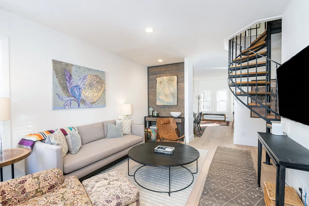 The image showcases a beautifully remodeled living area featuring a plush sofa, striking contemporary artwork, and a unique spiral staircase. The space feels warm and inviting, perfect for relaxing or gathering with friends and family after a day exploring Telluride.